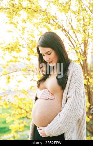 Außenporträt einer schönen schwangeren Frau, die den Bauch berührt und neben blühenden Frühlingsblumen posiert Stockfoto