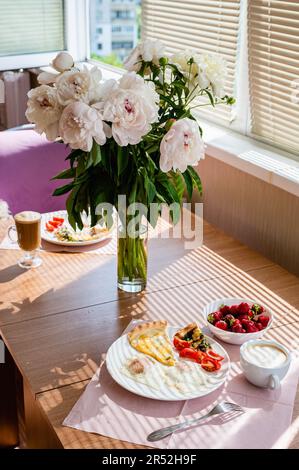 Ein wunderschöner Strauß Pfingstrosen steht in einer Vase auf dem Tisch. Frühstückskonzept. Spiegeleier mit einer Scheibe Pizza und einer Tasse Cappuccino auf dem Tisch Stockfoto