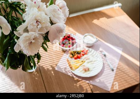 Spiegeleier mit einer Scheibe Pizza und einer Tasse Cappuccino auf dem Tisch. Ein wunderschöner Strauß Pfingstrosen steht in einer Vase auf dem Tisch. Frühstückskonzept Stockfoto