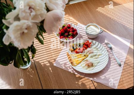 Spiegeleier mit einer Scheibe Pizza und einer Tasse Cappuccino auf dem Tisch. Ein wunderschöner Strauß Pfingstrosen steht in einer Vase auf dem Tisch. Frühstückskonzept Stockfoto