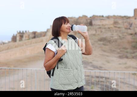 Erwachsene Reisende Frau mit Rucksack trinkt Wasser, während sie sich nach den Spaziergängen in den Ruinen der alten europäischen Festung im Freien entspannen. Rentner summieren sich Stockfoto