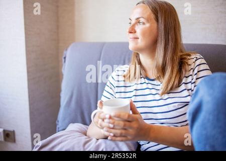 Porträt einer lächelnden Frau, die mit zwei Händen einen Becher hält, während sie sich auf dem Sofa ausruht Stockfoto