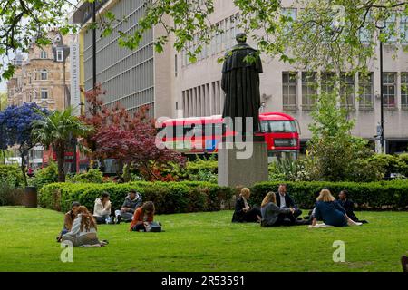 Cavendish Square London W1 England UK Sunshine mit Menschen, die auf Gras unter der Statue von Lord George Bentinck sitzen. Gebäude und Verkehr dahinter. Stockfoto