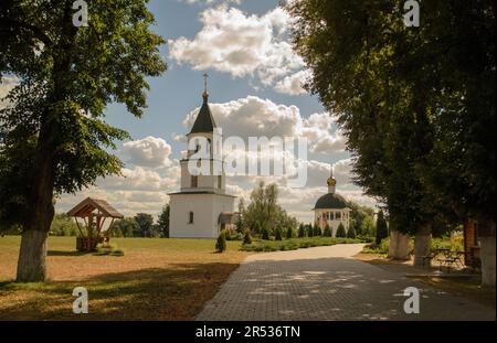 Die Pappelgasse führt zum weißen Glockenturm und weiter zum Gebetskreuz. Ein alter Holzbrunnen, auf der rechten Seite ist ein Pavillon im Schatten von Bäumen. Stockfoto