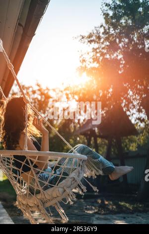 Glückliche junge Frau, die auf einem Makrame-Schaukelstuhl in der Nähe des Landhauses im Freien sitzt Stockfoto