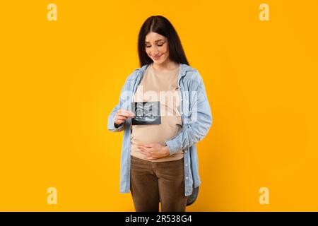 Aufgeregte junge Schwangere, die den Bauch umarmt und ein Ultraschallbild des ungeborenen Babys auf dem gelben Hinterkopf zeigt Stockfoto