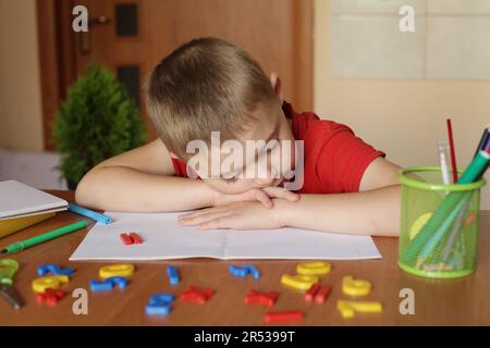 Der junge Junge liegt auf dem Tisch, macht ein Nickerchen beim Lernen und bereitet sich auf die Prüfung/Hausaufgaben vor. Erschöpftes und erschöpftes Schülerkonzept. Stockfoto