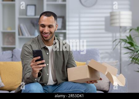 Ein zufriedener lateinamerikanischer Mann sitzt zu Hause auf dem Sofa und hält das Paket, das er per Kurier erhalten hat, auf. Verwendet das Telefon, nimmt eine Online-Bestellung über die Anwendung vor. Stockfoto