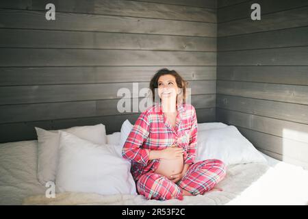 Morgenporträt einer glücklichen, jungen schwangeren Frau, die im Bett ruht und einen gemütlichen roten Pyjama trägt Stockfoto