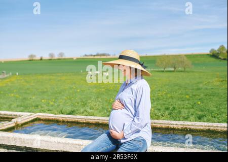 Außenporträt einer glücklichen jungen schwangeren Frau, die einen schönen Tag auf dem Land genießt, auf einem Brunnen sitzt, eine grüne Weide mit Kühen im Hintergrund Stockfoto