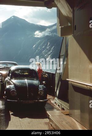 Geirangerfjord, Norwegen. Ca. 1960 – Eine junge Frau, die neben einem Volkswagen-Käfer auf dem Deck einer Autofähre auf dem Geirangerfjord steht. Stockfoto