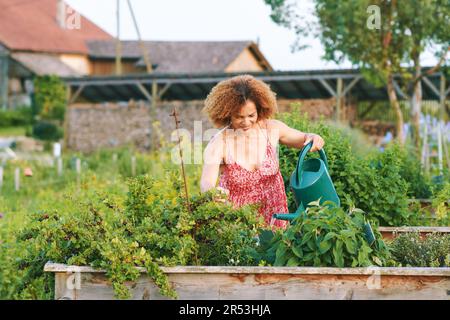 Außenporträt einer wunderschönen Frau mittleren Alters, die Pflanzen im Garten bewässert, rote Kleidung trägt, Glück und einen gesunden Lebensstil Stockfoto