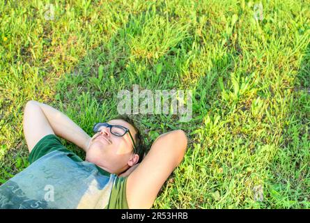 Ein junger Mann mit Brille lag auf dem Gras und genoss den Urlaub, nachdem er sehr glücklich im Park studiert hatte Stockfoto