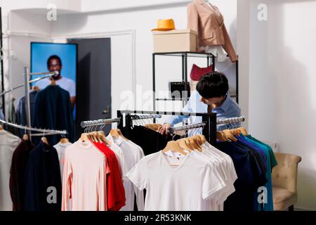 Ein eleganter Mitarbeiter, der im Geschäft arbeitet, organisiert visuelle Kleidung für die Eröffnung des Einkaufszentrums. Eine weiße Frau, die sich modische Ware anschaut, den Preis in einer modernen Boutique überprüft Stockfoto