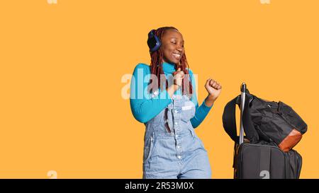 Ein fröhlicher Erwachsener hört Musik, tanzt und verwendet Kopfhörer, mit Trolley-Taschen und Gepäck vor der Kamera. Junger Reisender, der Tanzbewegungen zeigt und mp3 Songs auf dem Headset genießt. Stockfoto