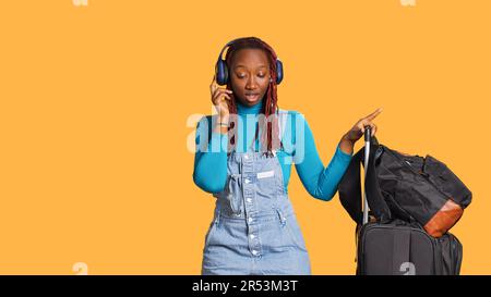 Fröhliches Mädchen, das Musik hört, tanzt und ein Audio-Headset benutzt, einen Trolley und Reisetaschen im Studio hat. Lächelnde Frau, die Tanzbewegungen macht und mp3 Songs über Kopfhörer hört. Stockfoto