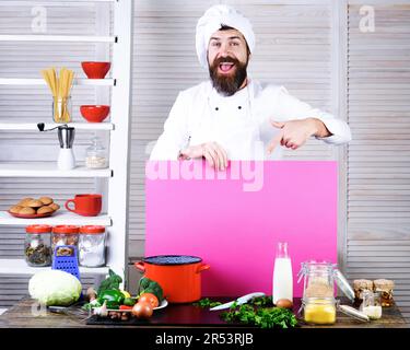 Der Koch mit dem glücklichen Bart zeigt mit dem Finger auf ein leeres Brett, um die Speisekarte anzupreisen. Koch in Uniform mit leerer Reklametafel für Werbetexte in Stockfoto