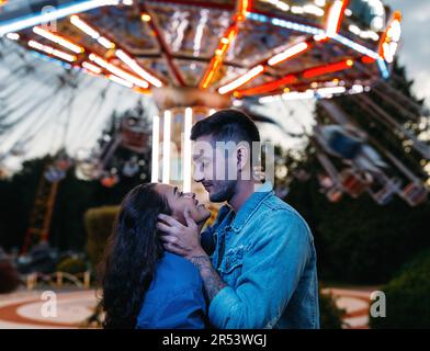 Glückliches Paar in einem Vergnügungspark. Ein junges Paar, das sich umarmt und sich ansieht, gegen das Karussell. Stockfoto