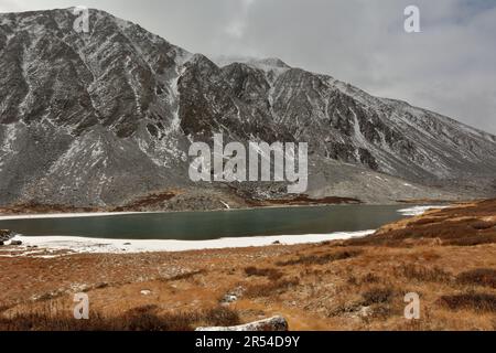 Das Ufer des Sees mit Eis am Wasserrand, überwuchert mit Gras und Moos auf dem sanften Hang der Bergkette, bestreut mit dem ersten Stockfoto