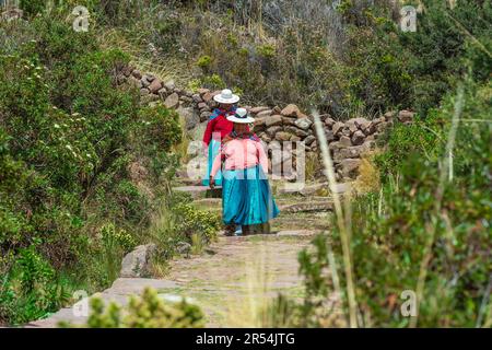 Zwei einheimische Frauen in Quechua in traditioneller Kleidung, die den Pfad zum Hafen von Isla Taquile (Taquile Island), Titicaca See, Peru hinunter gehen. Stockfoto