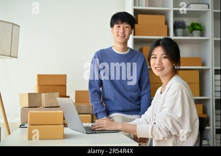 Ein glückliches und erfolgreiches junges asiatisches Paar, Inhaber eines kleinen Online-Unternehmens sind in ihrem Heimbüro, lächeln und schauen in die Kamera. Unternehmensgründung, Stockfoto