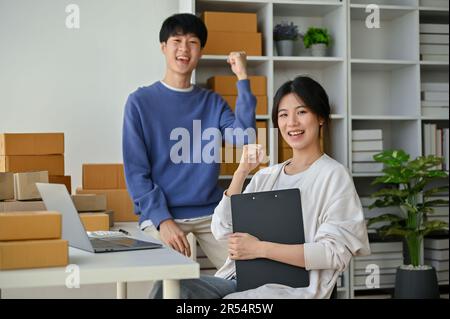 Glückliches und erfolgreiches junges asiatisches Paar, Inhaber eines KMU-Online-Unternehmens zeigen ihre Fäuste, während sie zusammen im Home Office arbeiten. Stockfoto