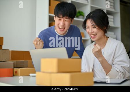 Ein fröhliches und erfolgreiches junges asiatisches Paar, die Inhaber von Online-Unternehmen zeigen ihre Fäuste und feiern gemeinsam ihren Erfolg im Online-Geschäft. KMU Stockfoto