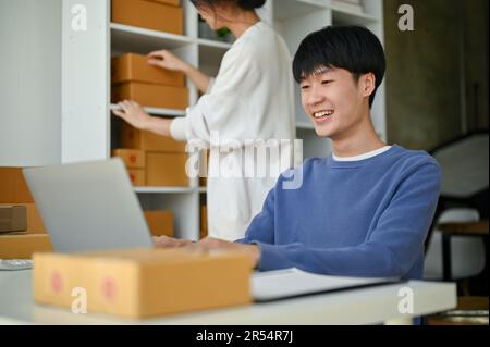 Ein glücklicher junger asiatischer, männlicher Online-Shop-Besitzer arbeitet in seinem Büro und überprüft Bestellungen auf der Website auf seinem Laptop, während seine Freundin nach dem Laden sucht Stockfoto