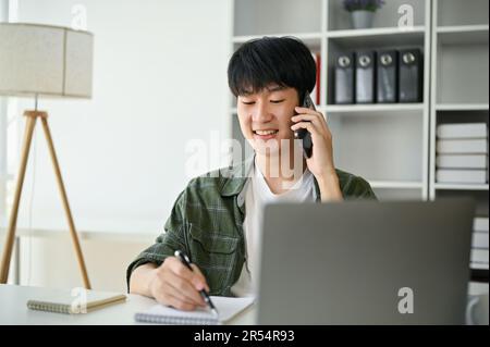 Ein glücklicher junger asiatischer Geschäftsmann oder Marketingassistent telefoniert mit einem Geschäftskunden an seinem Schreibtisch. Stockfoto