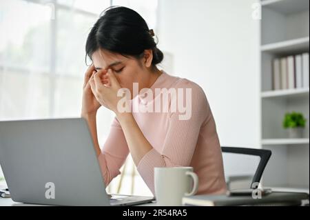 Gestresste und müde asiatische Geschäftsfrau sitzt an ihrem Schreibtisch in ihrem Büro und leidet unter Kopfschmerzen, während sie sich um ihre Arbeit sorgt. Stockfoto