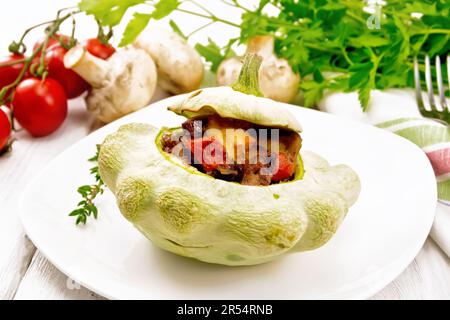 Patisson gefüllt mit Leber, Zwiebeln, Pilzen, Tomaten und Käse auf einem Teller, Handtuch, Champignons und Petersilie auf dem Hintergrund eines hellen Holzbretts Stockfoto