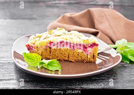 Ein Stück süßen Kuchen mit Himbeeren, Butterbröckeln und Mandelflocken auf einem Teller, Minze und Handtuch auf dunklem Holzbrettgrund Stockfoto