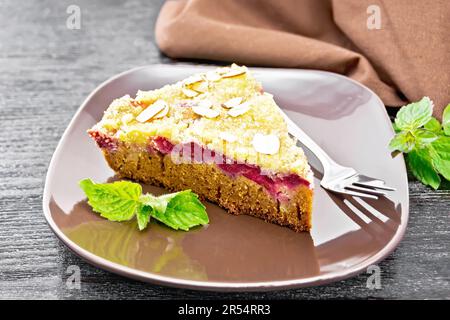 Ein Stück süßen Kuchen mit Himbeeren, Butterbröckeln und Mandelflocken auf einem Teller, Minze und Serviette auf dunklem Holzbrettgrund Stockfoto