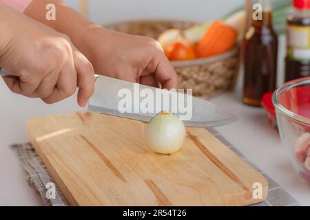 Weibliche Hand mit Messer, um weiße Zwiebeln auf Holzbrett zu schneiden. Eine Frau, die zu Hause in der Küche Essen zubereitet. Stockfoto