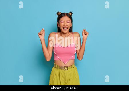 Thai Slim Girl in rosa Short Top steht auf blauem Hintergrund, schließt die Augen mit Freude und erhebt die Fäuste, fantastisches Nachrichtenkonzept, Kopierraum Stockfoto