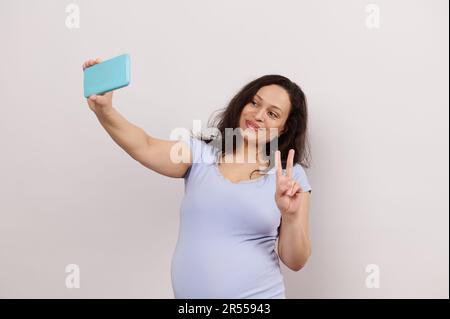 Wunderschöne, multiethnische, junge, schwangere Frau zeigt ein Friedensschild, Selfie auf dem Smartphone zu machen, spricht per Videoanruf Stockfoto