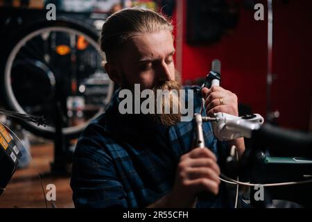 Nahaufnahme eines konzentrierten Fahrradmechanikers, der den Fahrradlenker mit Werkzeugen in der Fahrradreparaturwerkstatt im Dunkeln überprüft und repariert Stockfoto