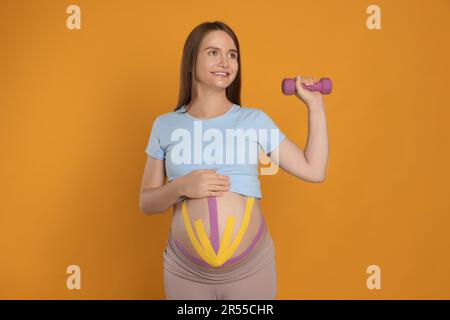 Schwangere Frau mit Kinesio-Bändern auf dem Bauch, die Übungen vor orangefarbenem Hintergrund macht Stockfoto