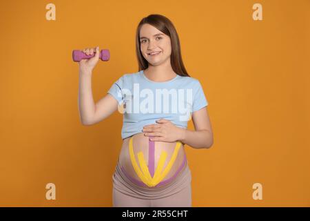 Schwangere Frau mit Kinesio-Bändern auf dem Bauch, die Übungen vor orangefarbenem Hintergrund macht Stockfoto