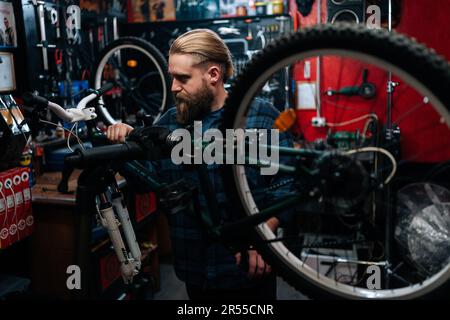Bärtiger Techniker, der Mountainbikes auf einem Fahrradständer repariert und in einer Fahrradwerkstatt mit dunklem Innenraum arbeitet. Männlicher Mechaniker, der Diagnosen durchführt Stockfoto