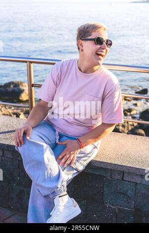 Androgyne nicht-binäre Person mit lebhaften kurzen Haaren, Piercings und sechseckiger Sonnenbrille, die am Meer lacht. Sommerliche Atmosphäre und wunderschöne Küste Stockfoto