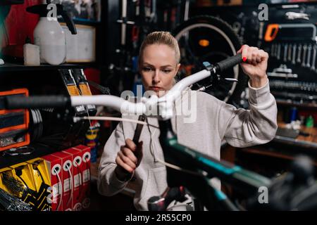 Konzentrierte blonde Fahrradmechanikerin, weibliche Befestigung des Fahrradlenkers mit Spezialwerkzeugen, die in einer Fahrradwerkstatt mit dunklem Innenraum arbeitet. Stockfoto