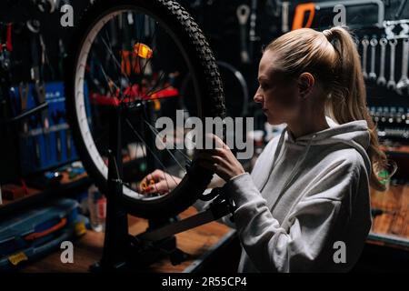 Vorderansicht einer fokussierten blonden, weiblichen Fahrradwerkstatt, die die Fahrradspeiche mit dem Fahrradspeichen-Schlüssel in einer Reparaturwerkstatt mit dunklem Innenraum überprüft. Stockfoto