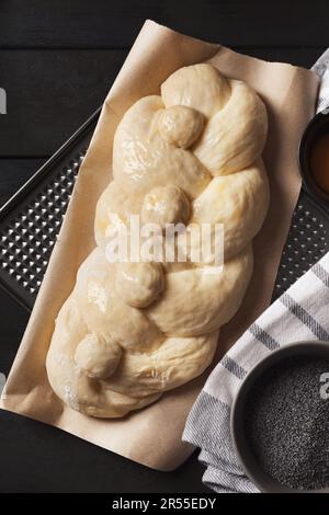 Rohes geflochtenes Brot und Mohnsamen auf schwarzem Holztisch, flach liegend. Traditionelle Sabbat-Challah Stockfoto