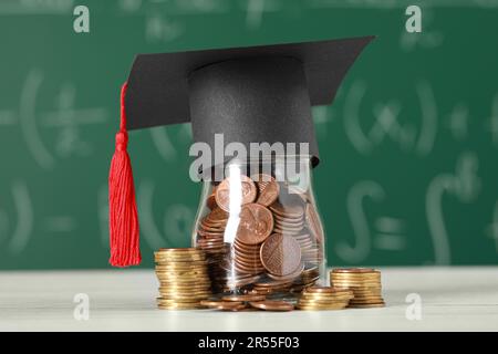 Stipendienkonzept. Glasbecher mit Münzen und Graduierungskappe auf weißem Holztisch Stockfoto