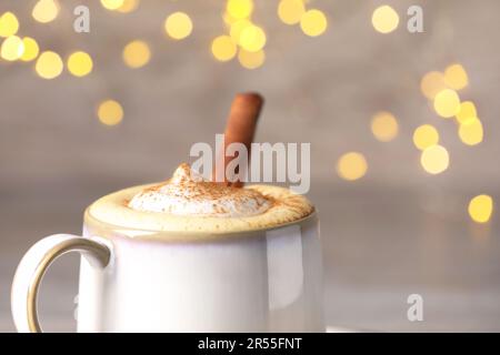 Tasse leckeren Eierpunsch mit Zimt auf verschwommenem Hintergrund vor festlichen Lichtern, Nahaufnahme Stockfoto