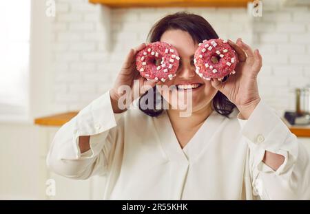 Nahaufnahme eines fröhlichen, lächelnden, dicken, dicken Weibes, das Spaß mit Donut-Kuchen auf den Augen hat. Stockfoto