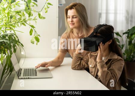 Familien und neue Technologien. Lächelnde Arbeit-zu-Hause-Mutter, die mit ihrer Tochter einen VR-Helm trägt und die virtuelle Realität erforscht. Neugieriges Teenager-Mädchen Stockfoto