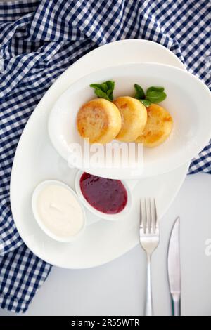 Runde Pfannkuchen mit Hüttenkäse auf einem weißen Teller auf blauem Hintergrund. Essen Frühstück. Gesundes Essen. Saure Sahne Sauce und Minze. Draufsicht Nahaufnahme. Dessertmahlzeit. Ricotta-Pommes aus Quark und Käse. Stockfoto