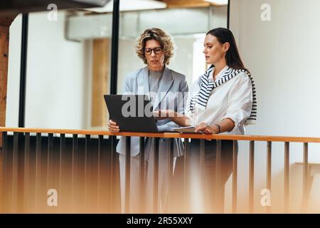 Zwei Geschäftsfrauen diskutieren miteinander, während sie einen Laptop benutzen. Berufsfrauen analysieren ihren Arbeitsfortschritt in einem Stand-up-Meeting. Geschäftsfrauen Stockfoto